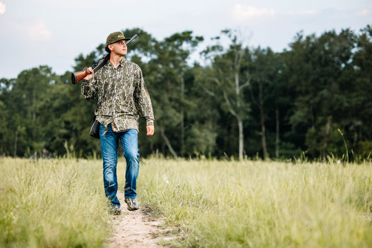 Man in a field wearing a Long-Sleeved Flyweight Camo Hunting Shirt, holding a gun, demonstrating the shirt's functional design and performance features.