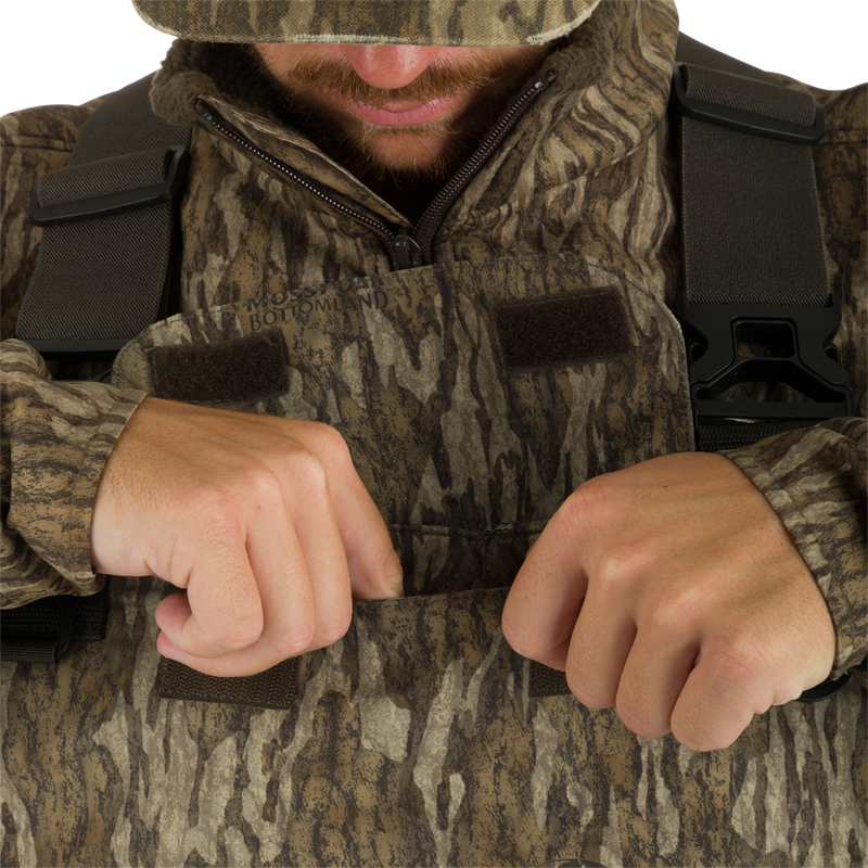 A man in camouflage waders with hands in pockets, showcasing the Insulated Guardian Elite Vanguard Breathable Waders by Drake Waterfowl. Waterproof, windproof, with LokDown insulation and Thinsulate boots for hunting comfort.