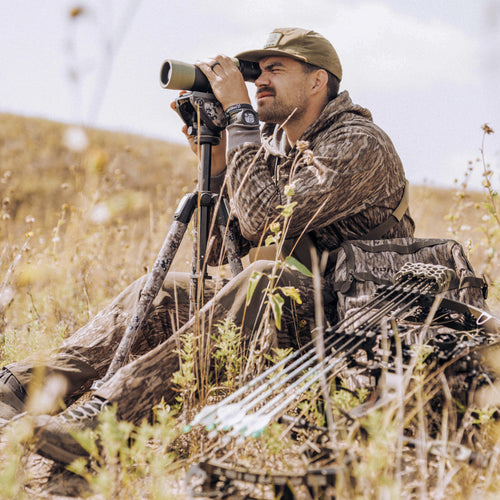 Man in camouflage using telescope, wearing Drake X Lost Hat DDTF Goat Rope Cap with flat visor and adjustable snap-back, embodying hunting gear aesthetics.