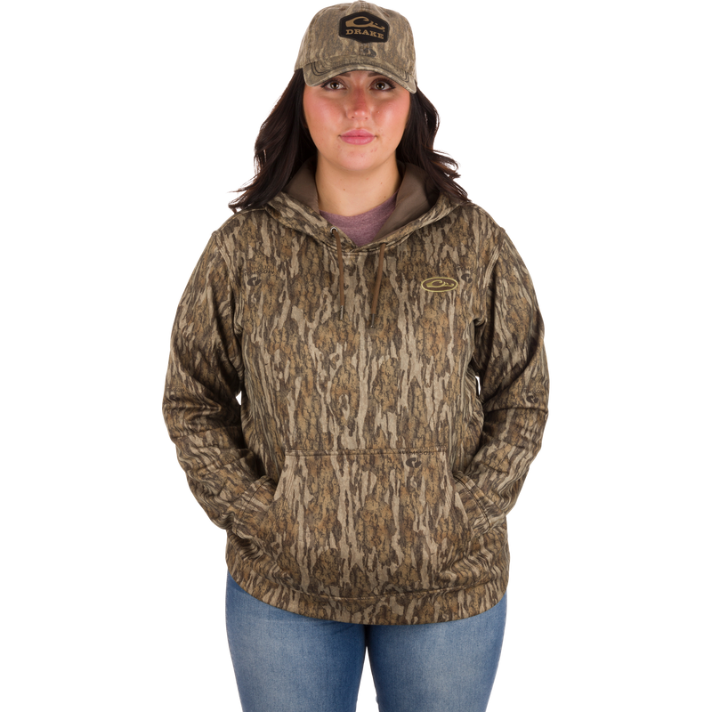 Woman in camouflage hoodie and hat wearing the Women's MST Performance Hoodie, featuring a double-lined hood, kangaroo pouch, and soft, combed fleece interior for warmth and comfort.