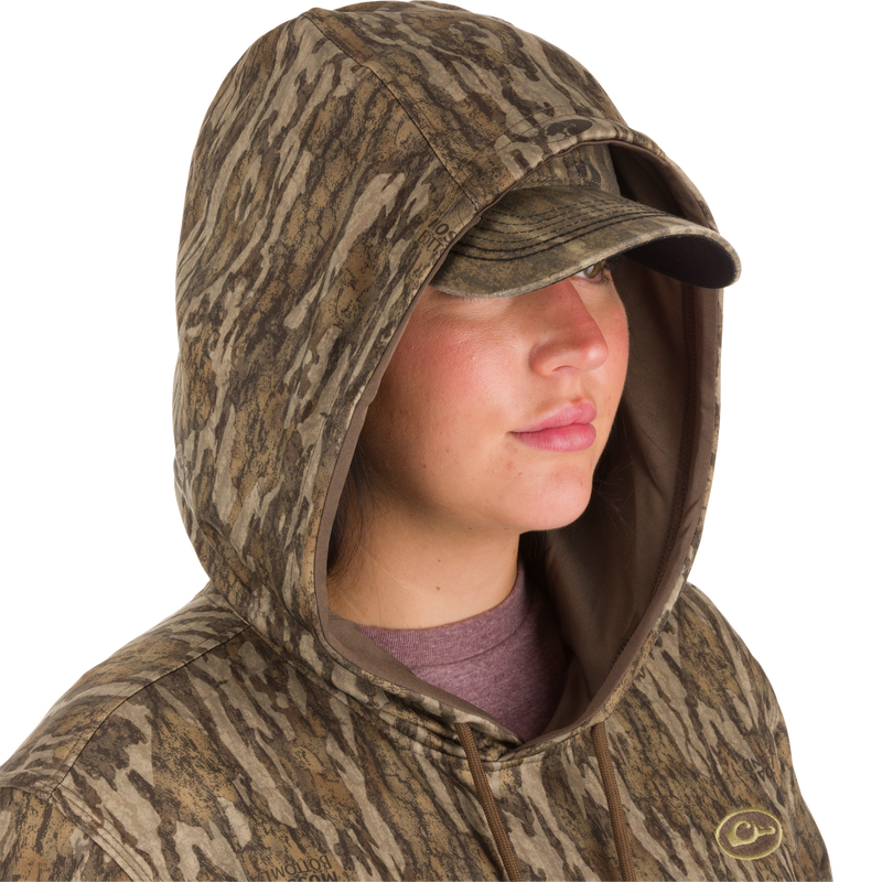 A woman wearing a Women's MST Performance Hoodie in camouflage with a visor, showcasing the hood and kangaroo pouch for enhanced comfort and wind protection.