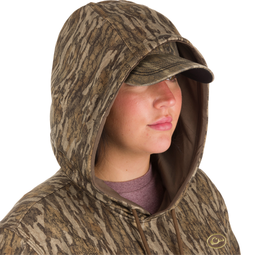 A woman wearing a Women's MST Performance Hoodie in camouflage with a visor, showcasing the hood and kangaroo pouch for enhanced comfort and wind protection.