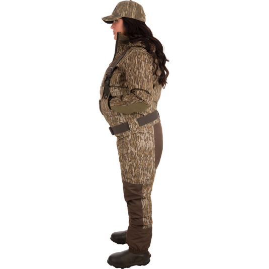 Woman wearing Women’s Insulated Guardian Elite Vanguard Breathable Waders with camouflage outfit, featuring reinforced seams, handwarmer pockets, front cargo pouch, and Thinsulate Buckshot Mud boots.