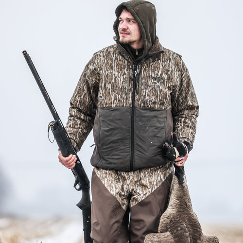 A man in a hooded jacket, the LST Guardian Flex Double Down Eqwader Full Zip, is holding a rifle in a snowy field.