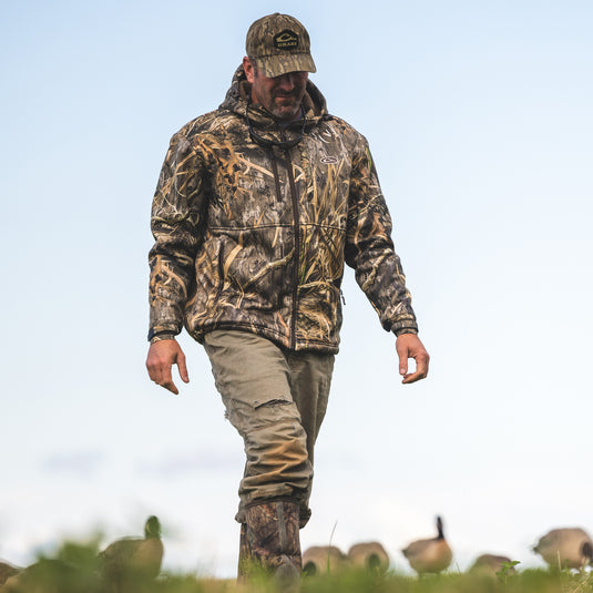 Man walking in a field, wearing the MST Hole Shot Hooded Windproof Eqwader Full Zip Jacket, featuring high handwarmer pockets, zippered lower slash pockets, and adjustable hood and waist.