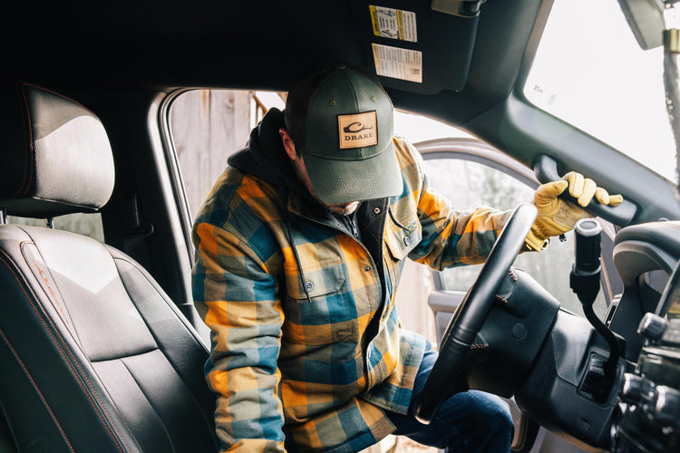 Man wearing The Drake Campfire Flannel Hoodie, driving a car, highlighting its warm, durable design with adjustable hood and button snap closure.