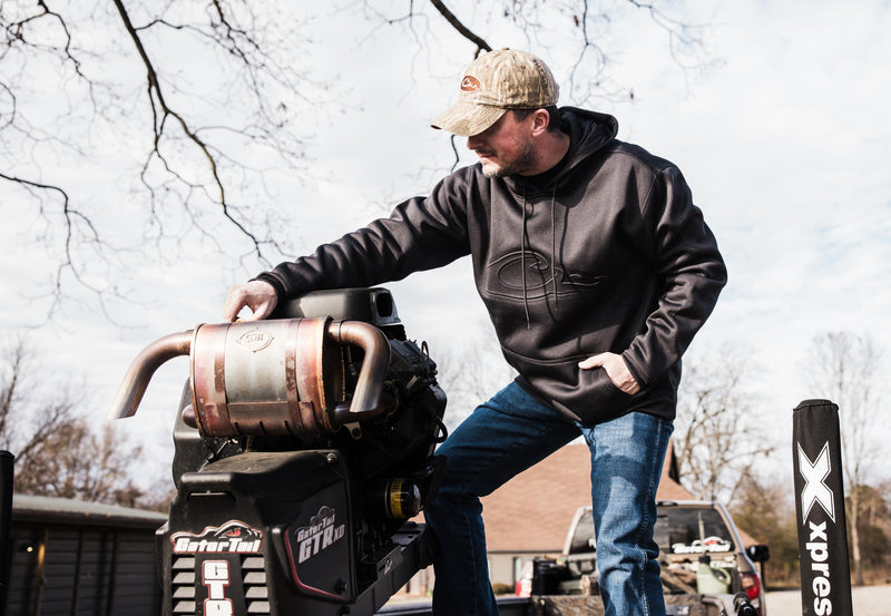 Man standing on a machine wearing the Drake Back Eddy Embossed Logo Hoodie, featuring a raised logo, kangaroo pocket, and adjustable hood with drawstrings.