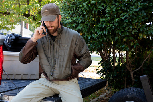 Man sitting on a truck, wearing the Two-Tone Synthetic Double-Down Full Zip jacket, talking on a cell phone, highlighting its versatile design for hunting and outdoor activities.