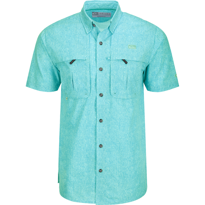 Heritage Heather Shirt S/S: A close-up of a blue shirt with pockets, featuring a logo. Lightweight, moisture-wicking, and quick-drying fabric for comfort. Hidden button-down collar and vented cape back for ventilation. Perfect for outdoor activities or office wear.